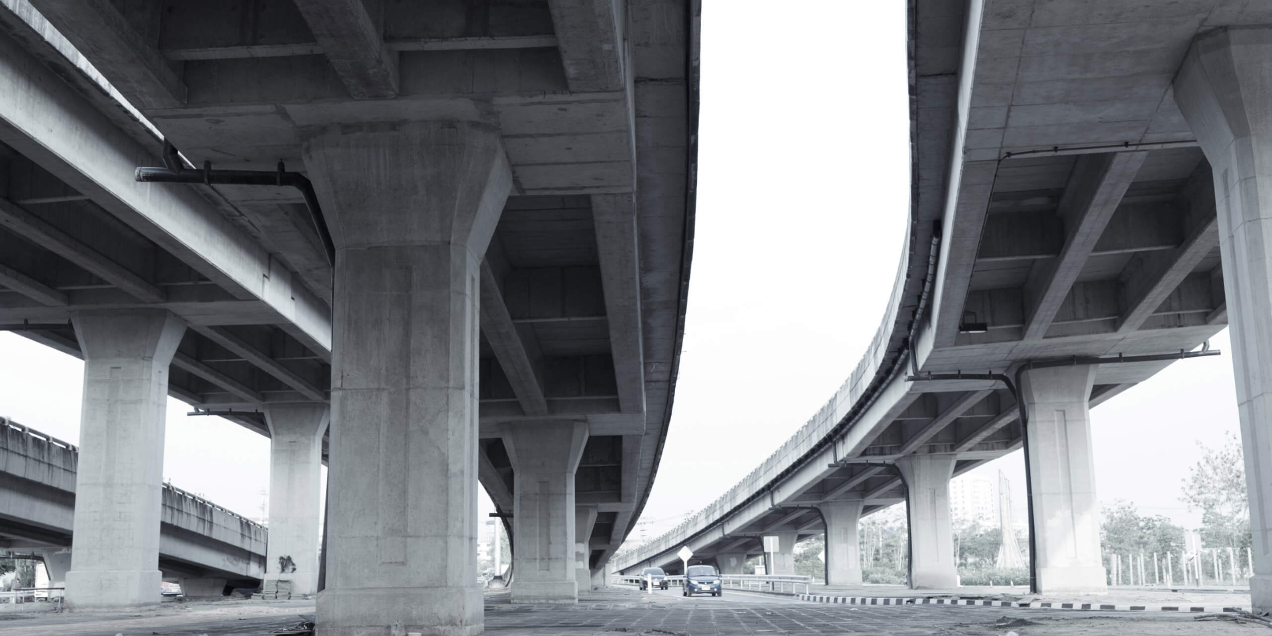 A view from under a bridge. Two cars visible in the distance. The curb on the right side is painted in black and white stripes.